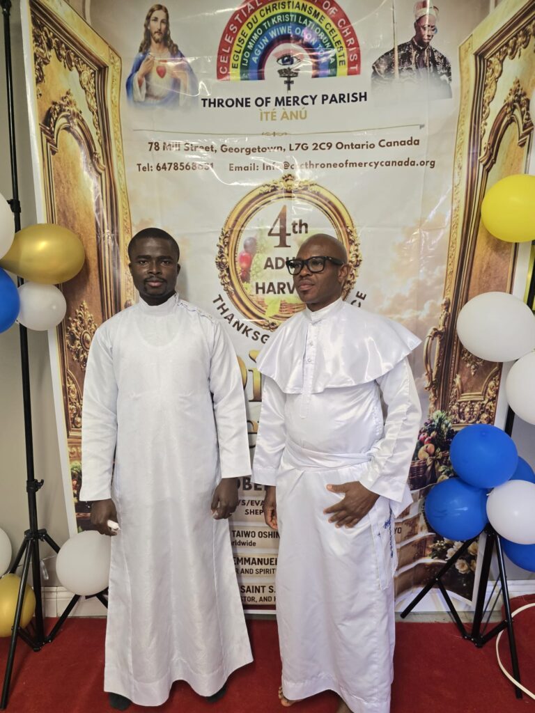 Two clergy members in white garments in front of the harvest thanksgiving banner